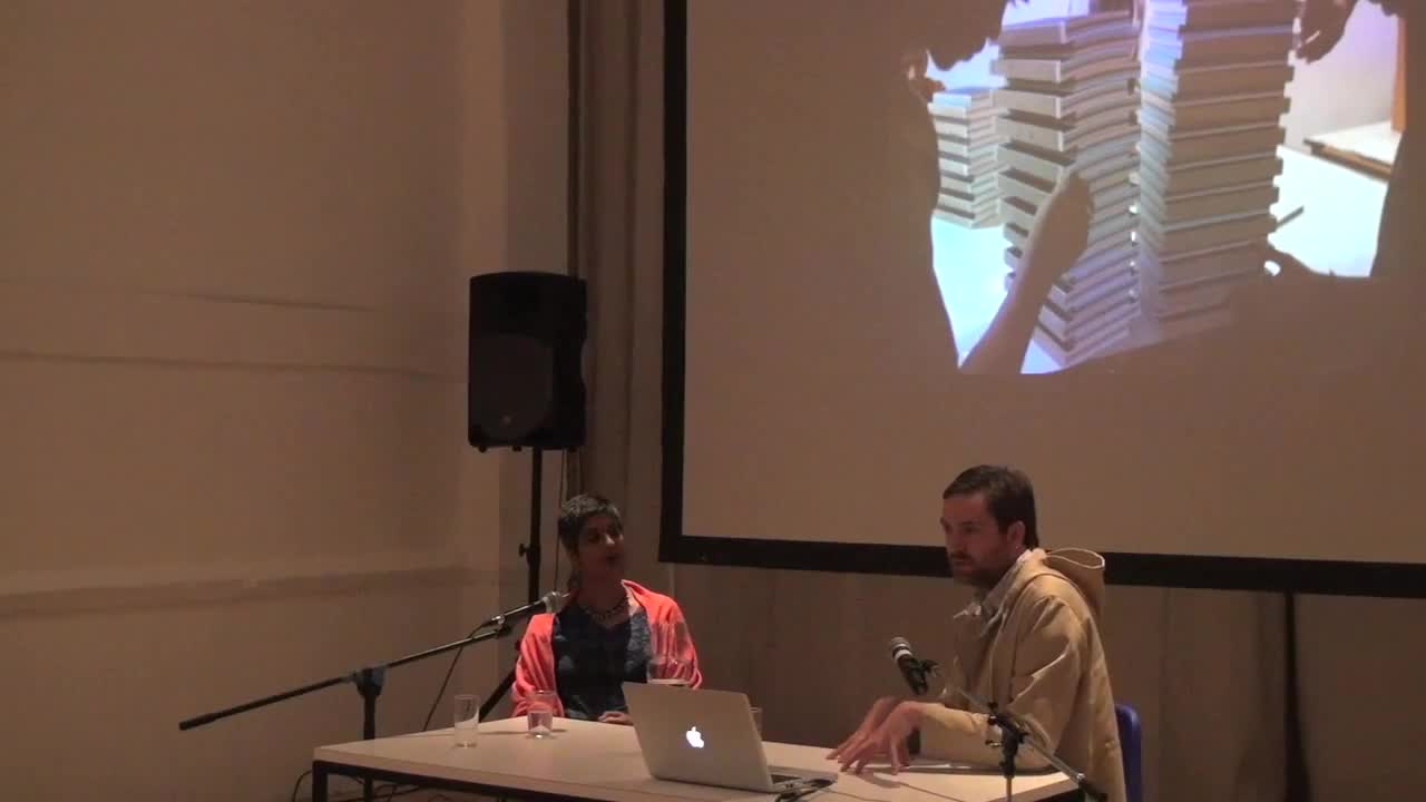 Video documentation of a man and woman seated at a table, giving a talk with a PowerPoint presentation projected on a screen behind them.