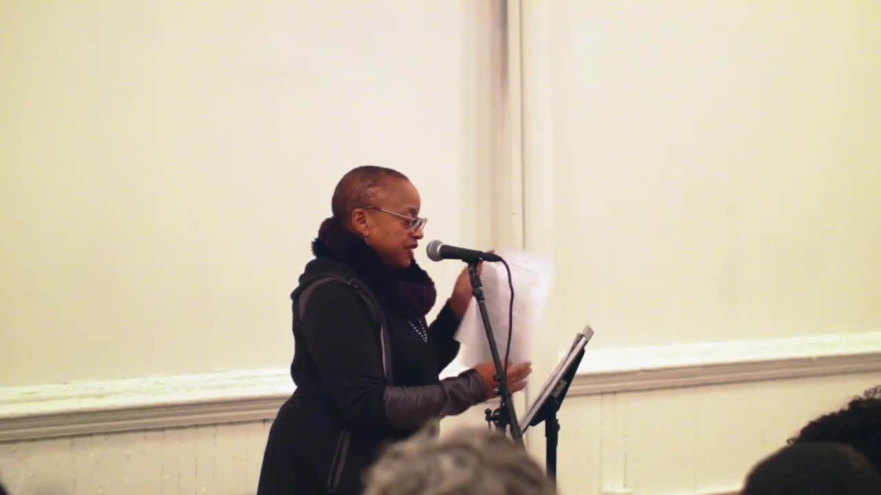A person reads from papers on a music stand, speaking to an audience with a microphone.