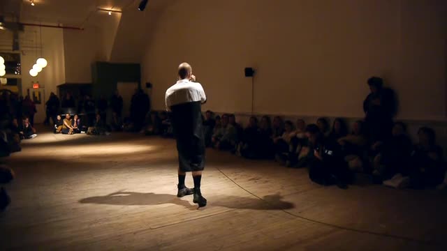 Video documentation of performers moving in the middle of a spotlit room, with the audience seated in a rectangle around them.