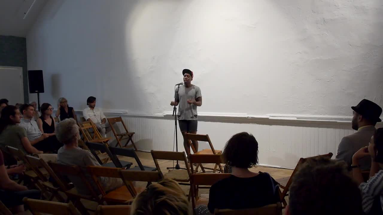 Video documentation of a man performing a reading for a seated audience.