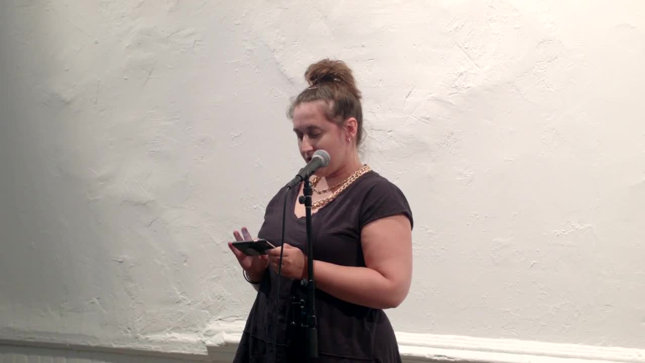 Video documentation of a woman performing a reading for a seated audience.