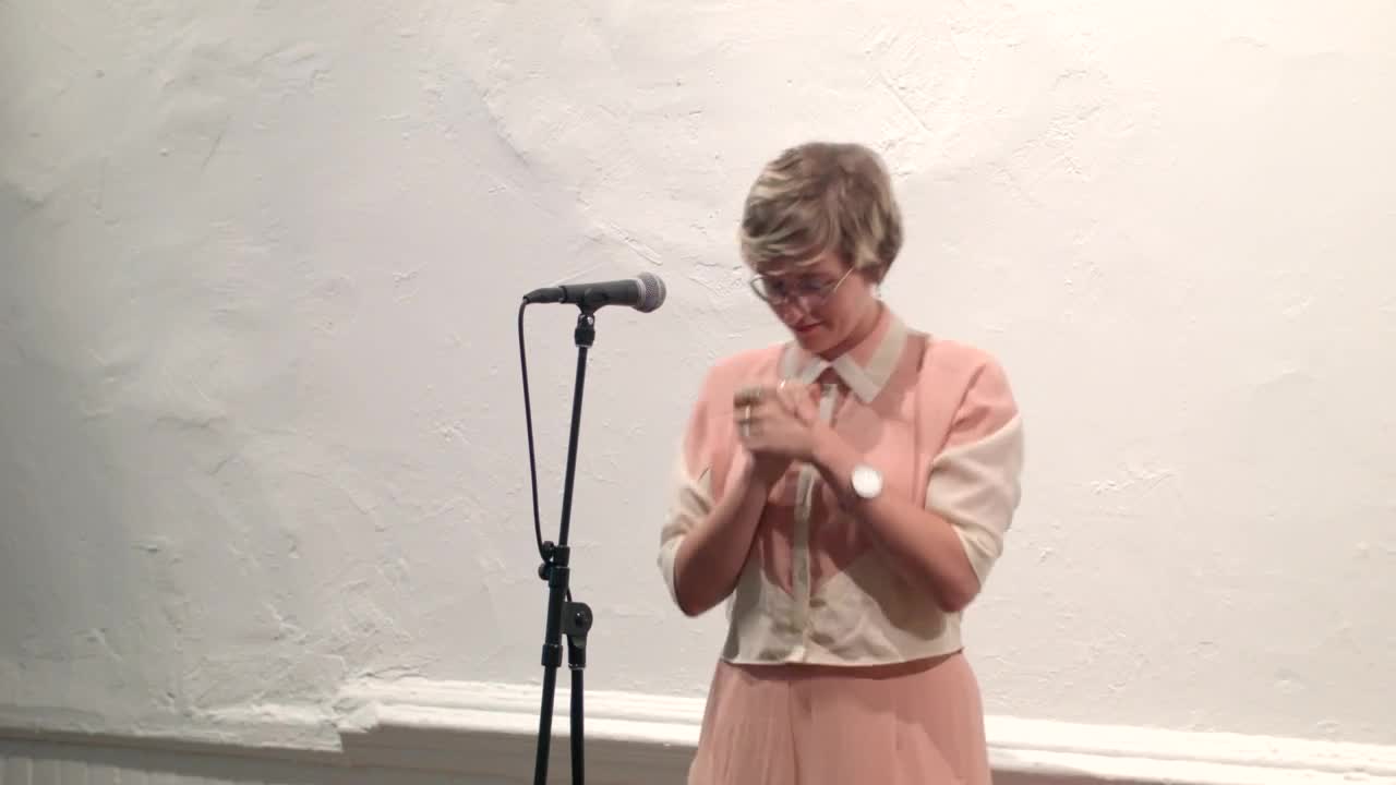 Video documentation of a woman performing a reading for a seated audience.