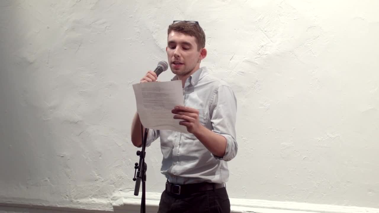 Video documentation of a man performing a reading for a seated audience.