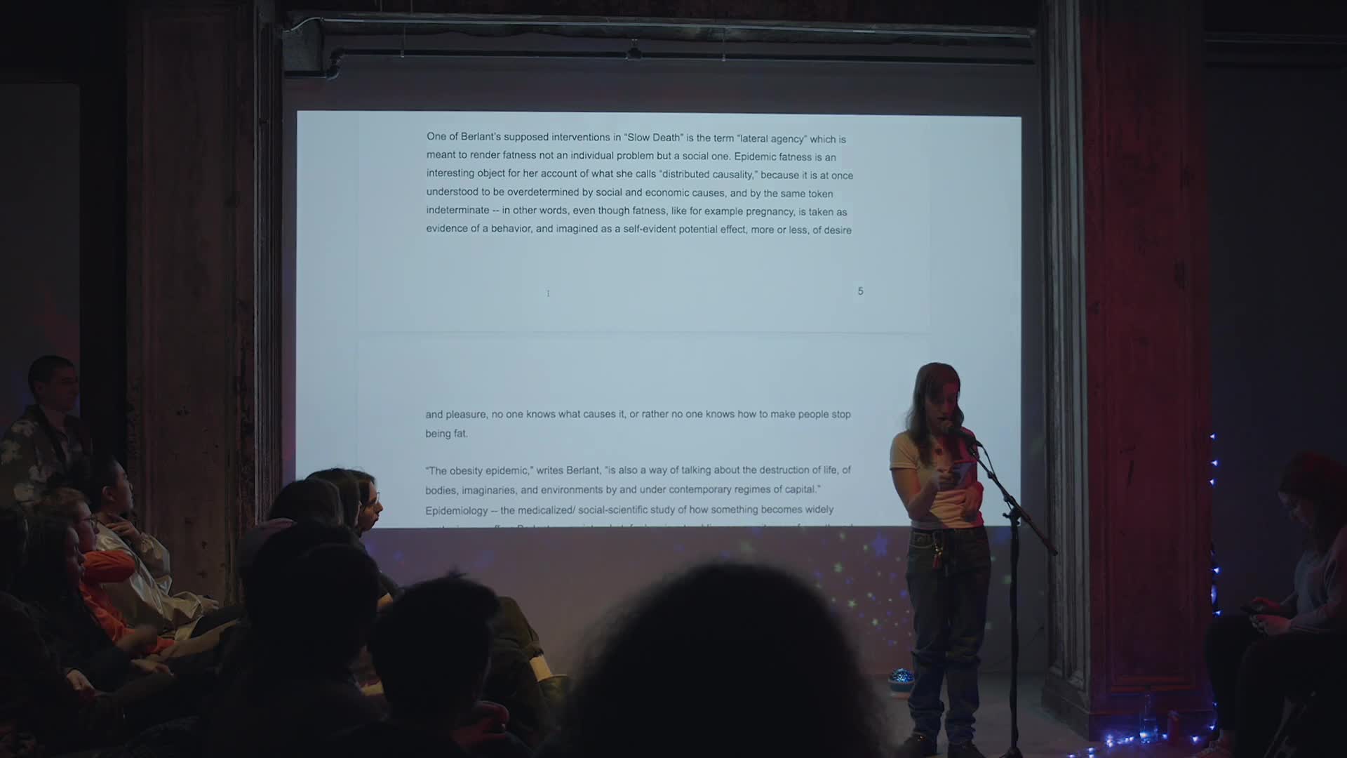 A person in a t-shirt and jeans stands at a microphone in front of a seated audience. She is reading off of her phone. There is a screen projected behind her that shows written text, videos, and photographs, changing in correspondence with what she is reading.