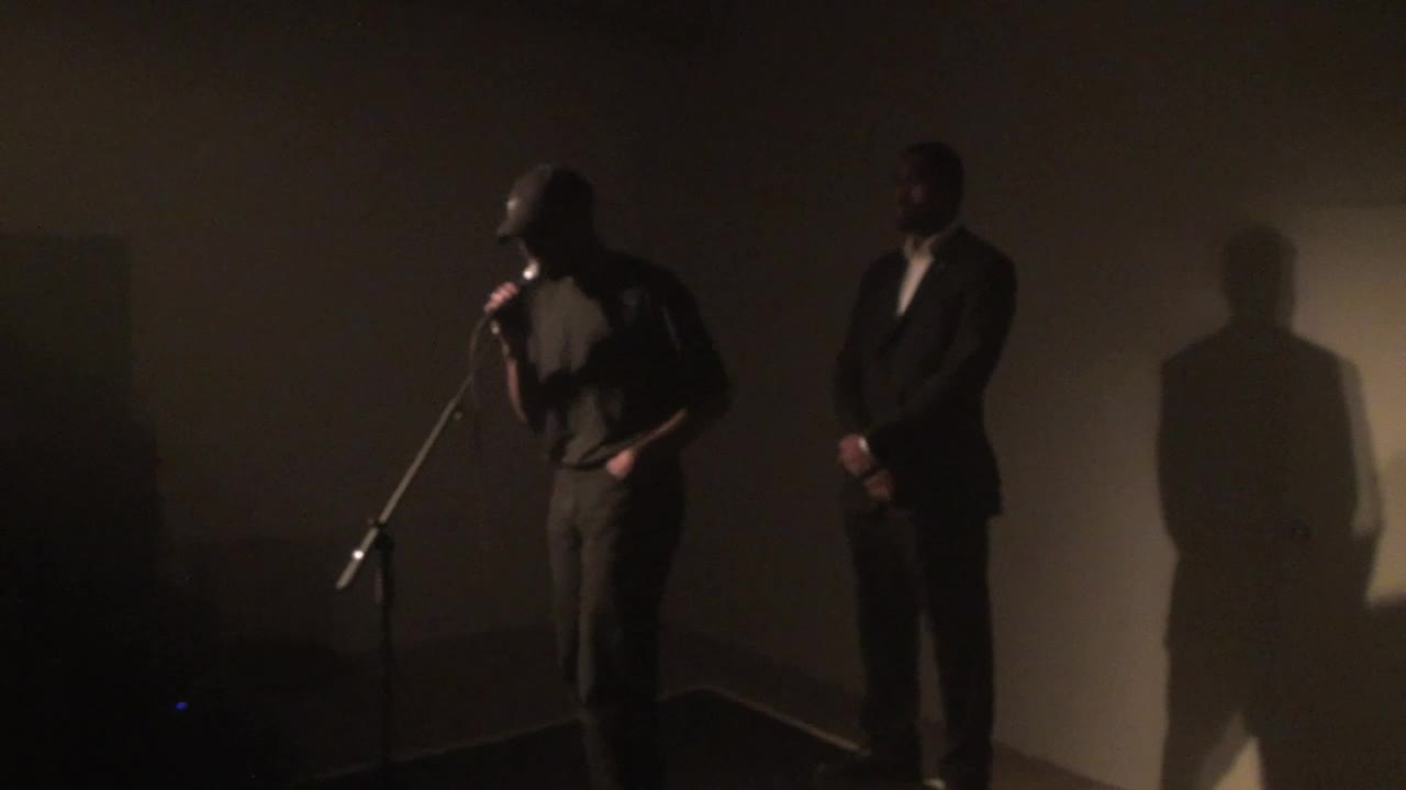Video documentation of a performance by a man dressed in black clothing and a hat.