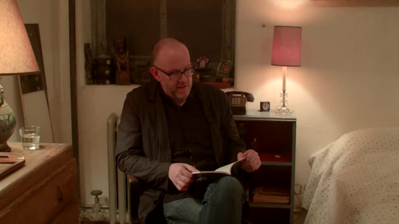 A man sits on a chair and reads from a small book in a bedroom.