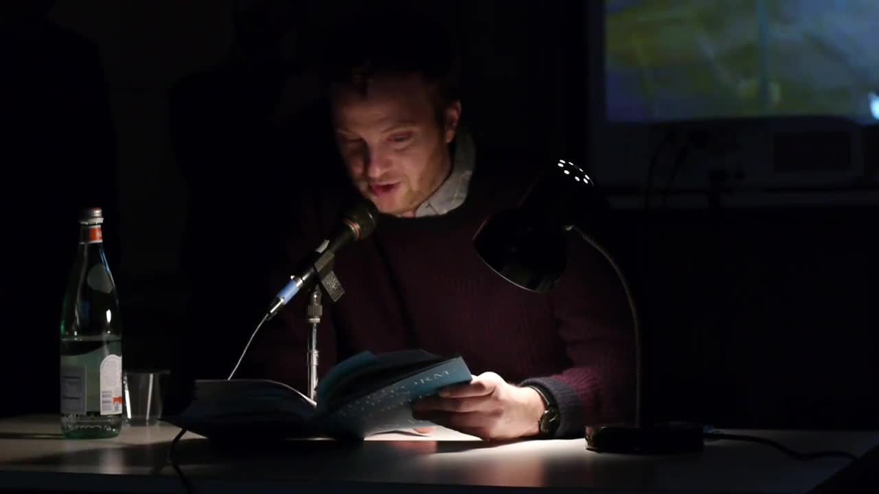 A person reads from a book illuminated by a lamp on a table in a dark room.