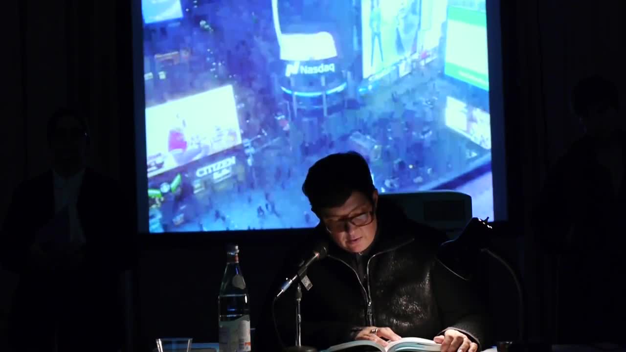 A person reads from a book illuminated by a lamp on a table in a dark room, with a video depicting an aerial view of Times Square projected in the background.