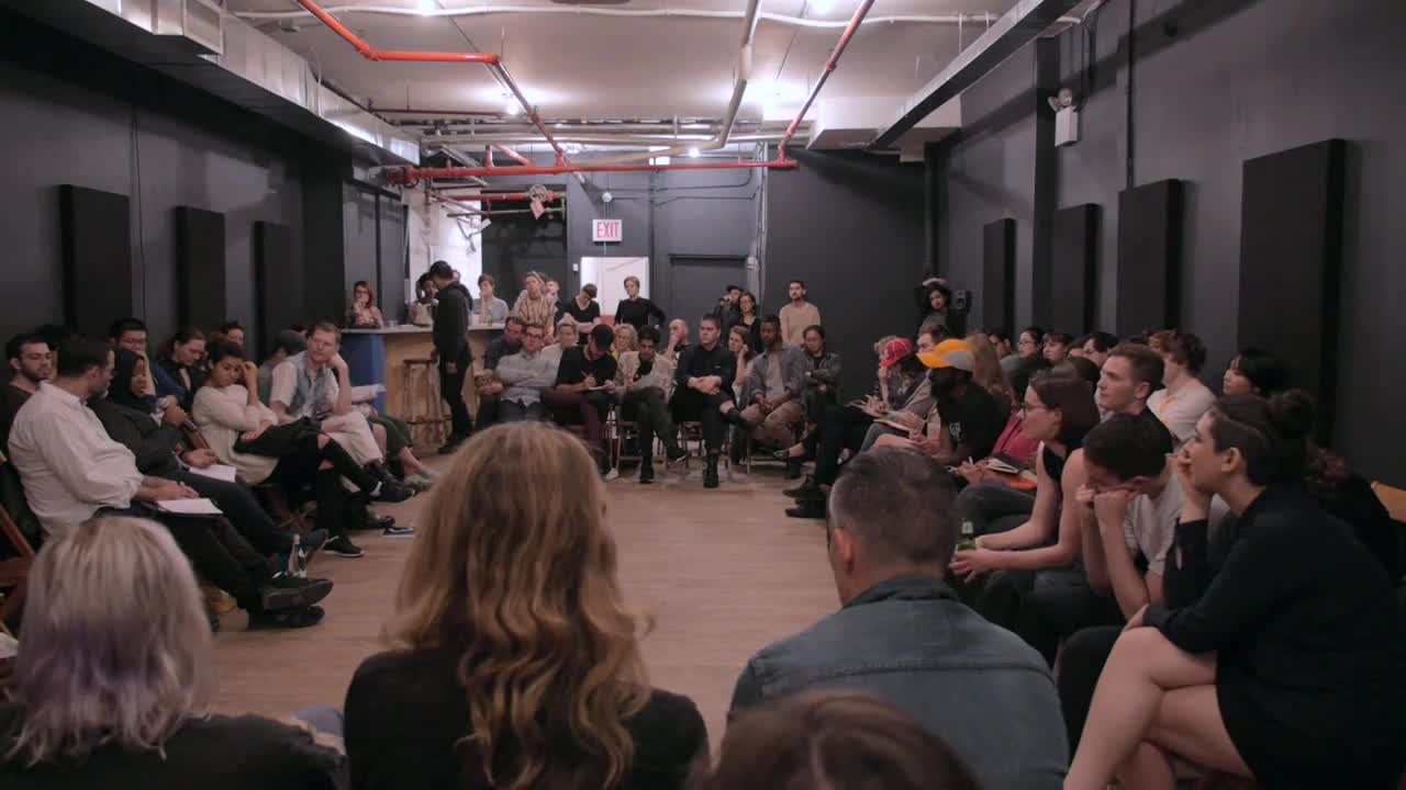 Several people speak on microphones among a crowd sitting in a circle around a room.