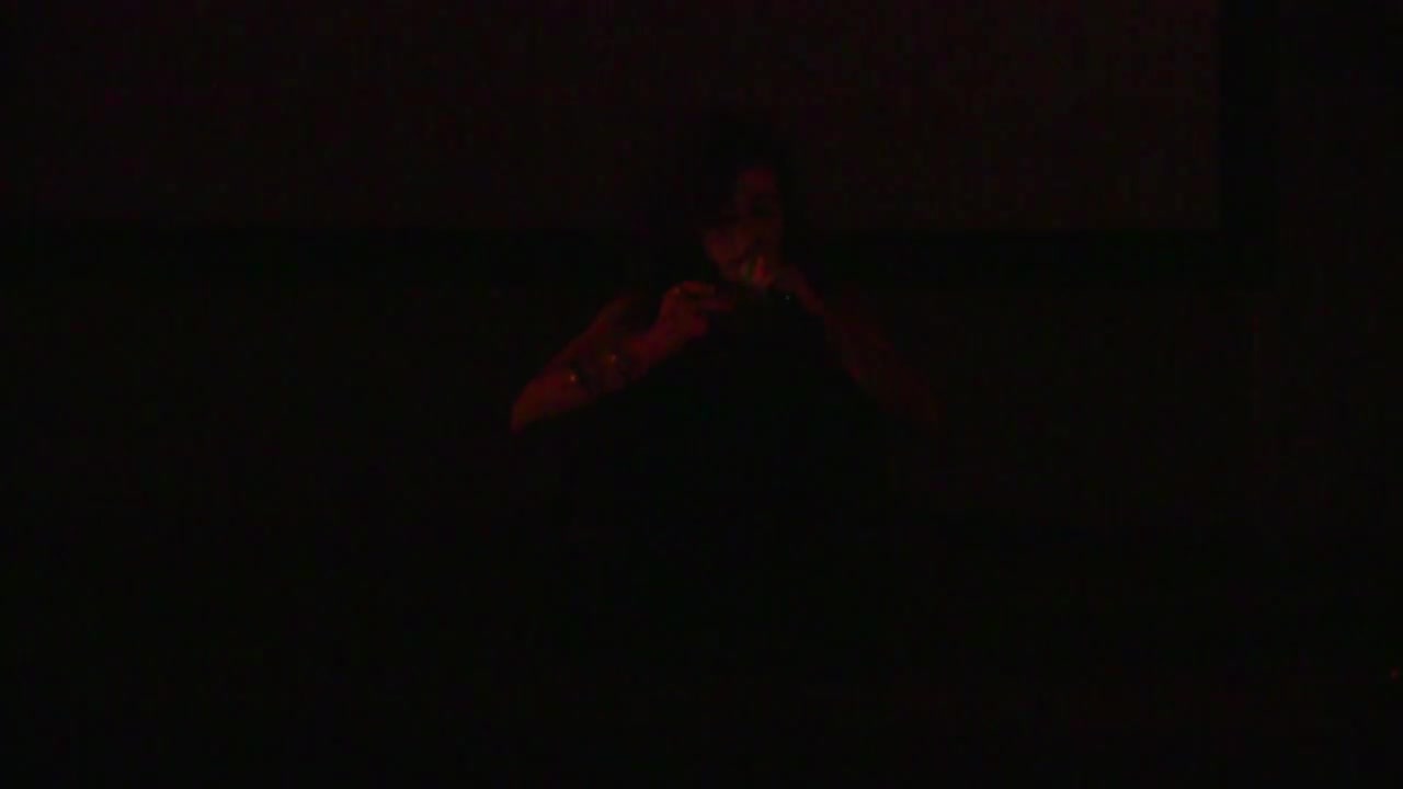 A figure illuminated by a red light in an otherwise dark space speaks into a microphone as they read a text.
