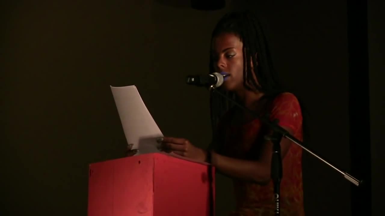 A figure sits behind a red podium and speaks into a microphone as they read from a piece of paper.