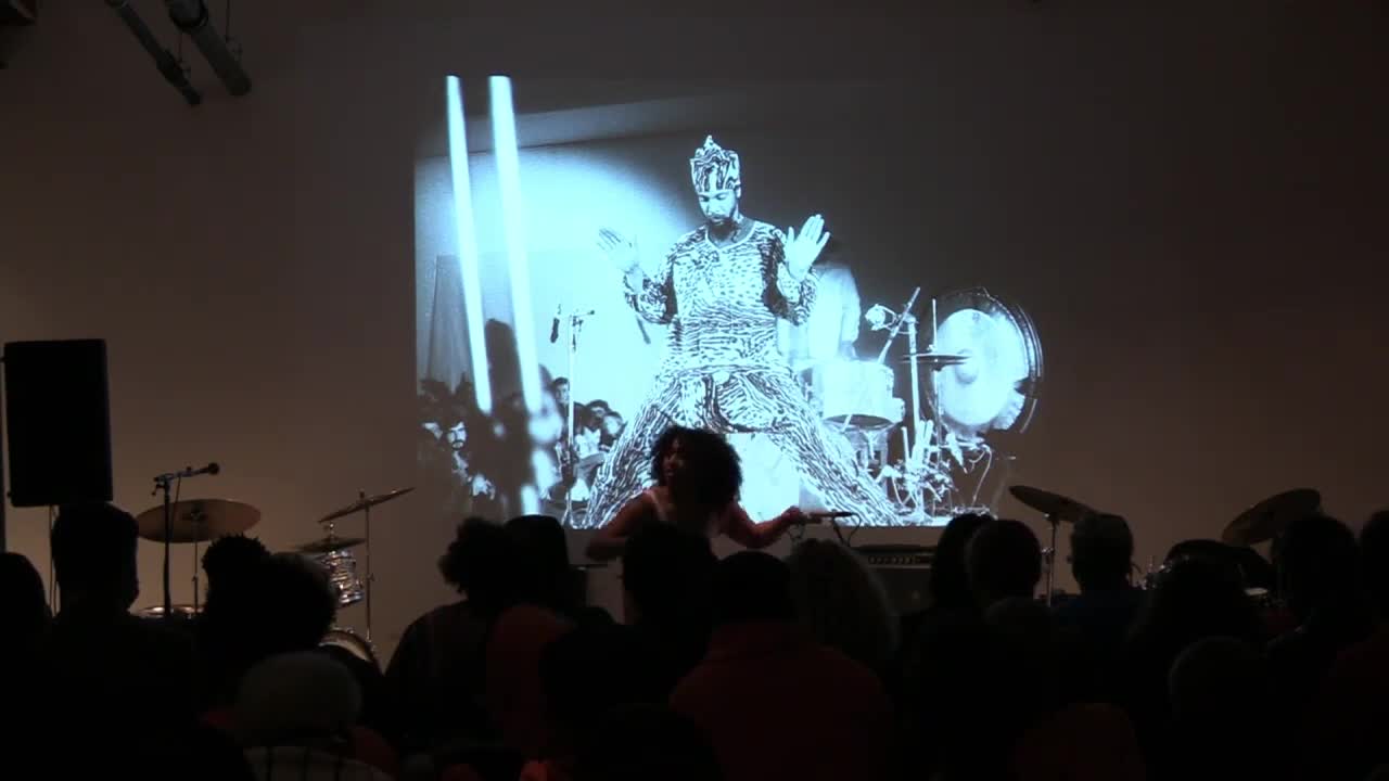 A color video of various figures performing, speaking, and moving in a basement space as a seated audience looks on. Behind the performers are projected images.