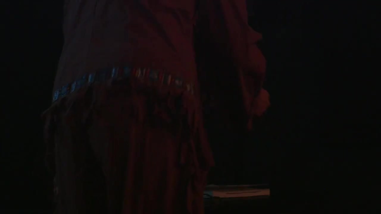 Performance in front of and among an audience, whereupon a man undresses and redresses in Native American clothing, using a microwave as a prop.
