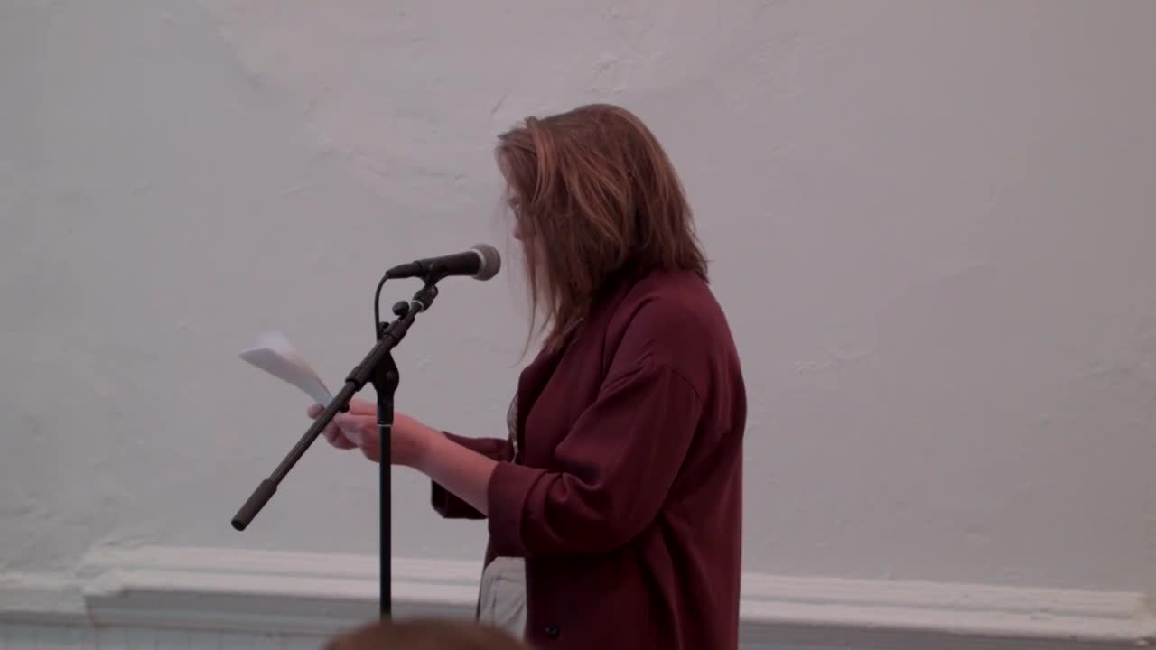 A person reads from a sheet of paper in front of a seated audience.
