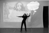 A black-and-white image of a woman, wearing all black clothing and sunglasses, standing with her arms raised. An image is projected on the wall behind her, with a description reading  