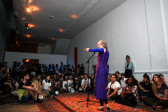 A woman stands in the middle of a room, surrounded by a crowd of people. She is speaking on a microphone, gesturing with her left arm, and standing on a carpet covering a raised section of the floor.