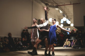 A photo of three performers standing in the middle of a room, surrounded by the audience. Two performers linked hands, and the third stands in between them, arms raised.