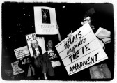 A black-and-white photo of a group of protestors holding various signs. One asks, 