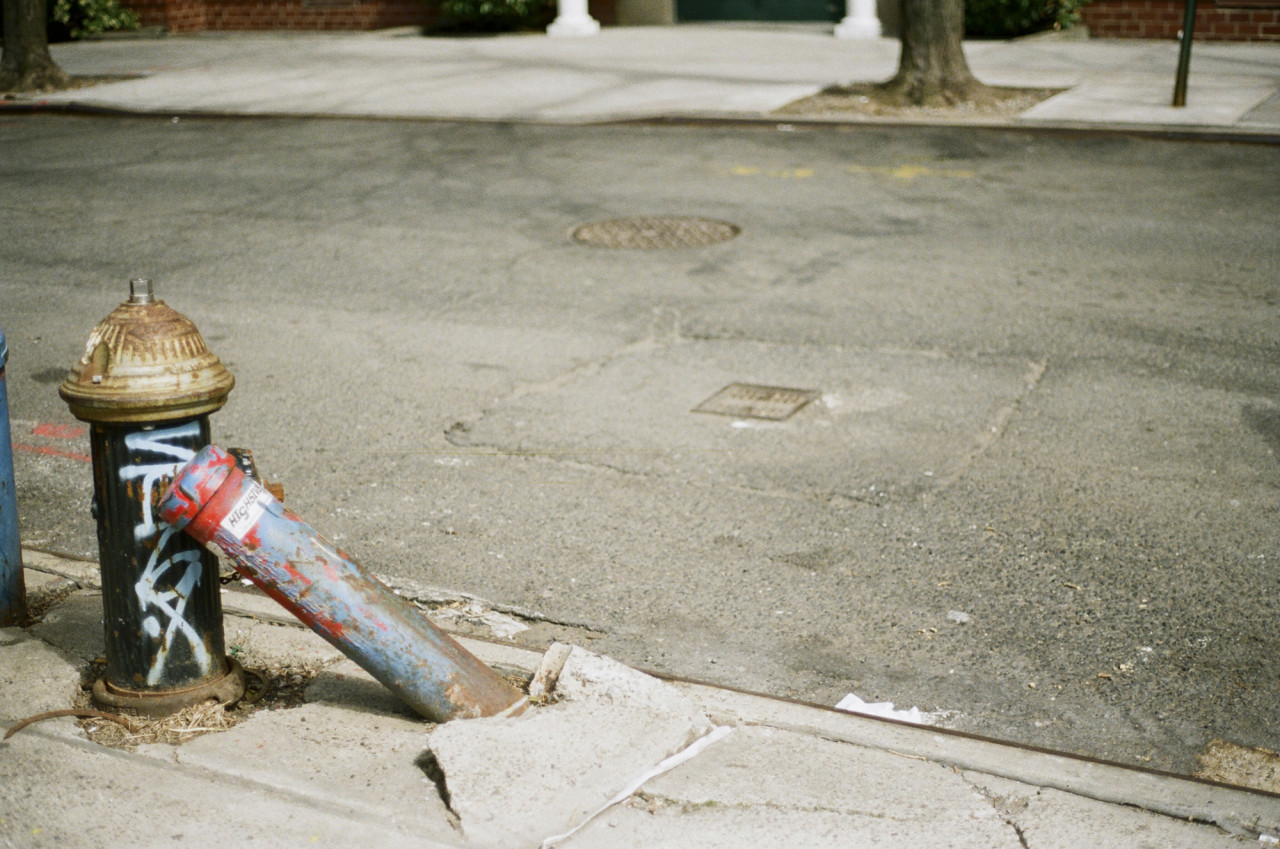 A photograph depicting a street in New York. A fire hydrant with some graffiti supports a leaning concrete pole, which has fallen over, lifting up a piece of the sidewalk