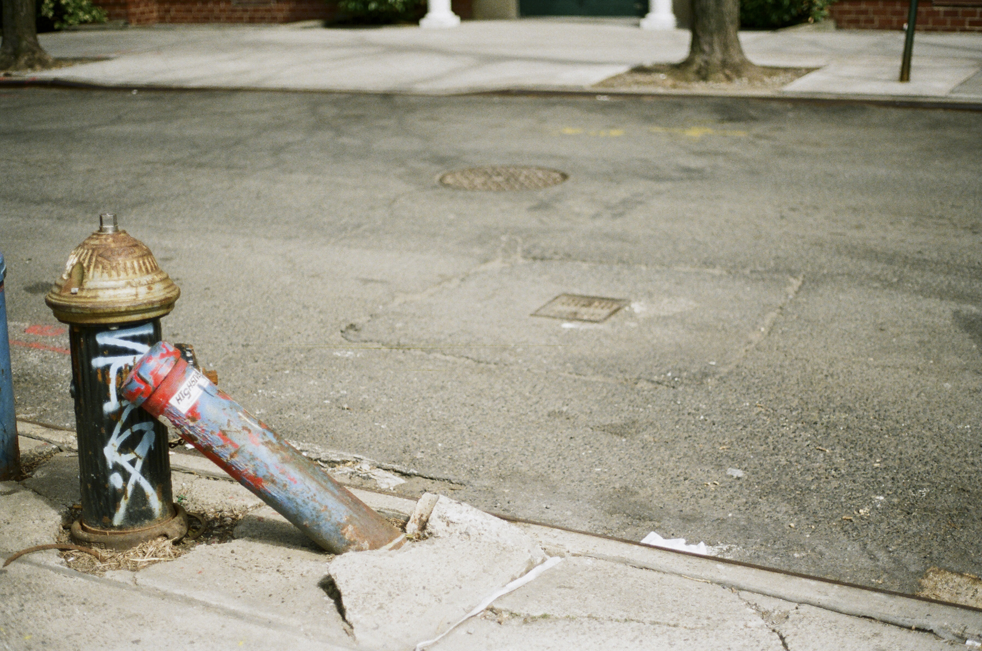 A photograph depicting a street in New York. A fire hydrant with some graffiti supports a leaning concrete pole, which has fallen over, lifting up a piece of the sidewalk