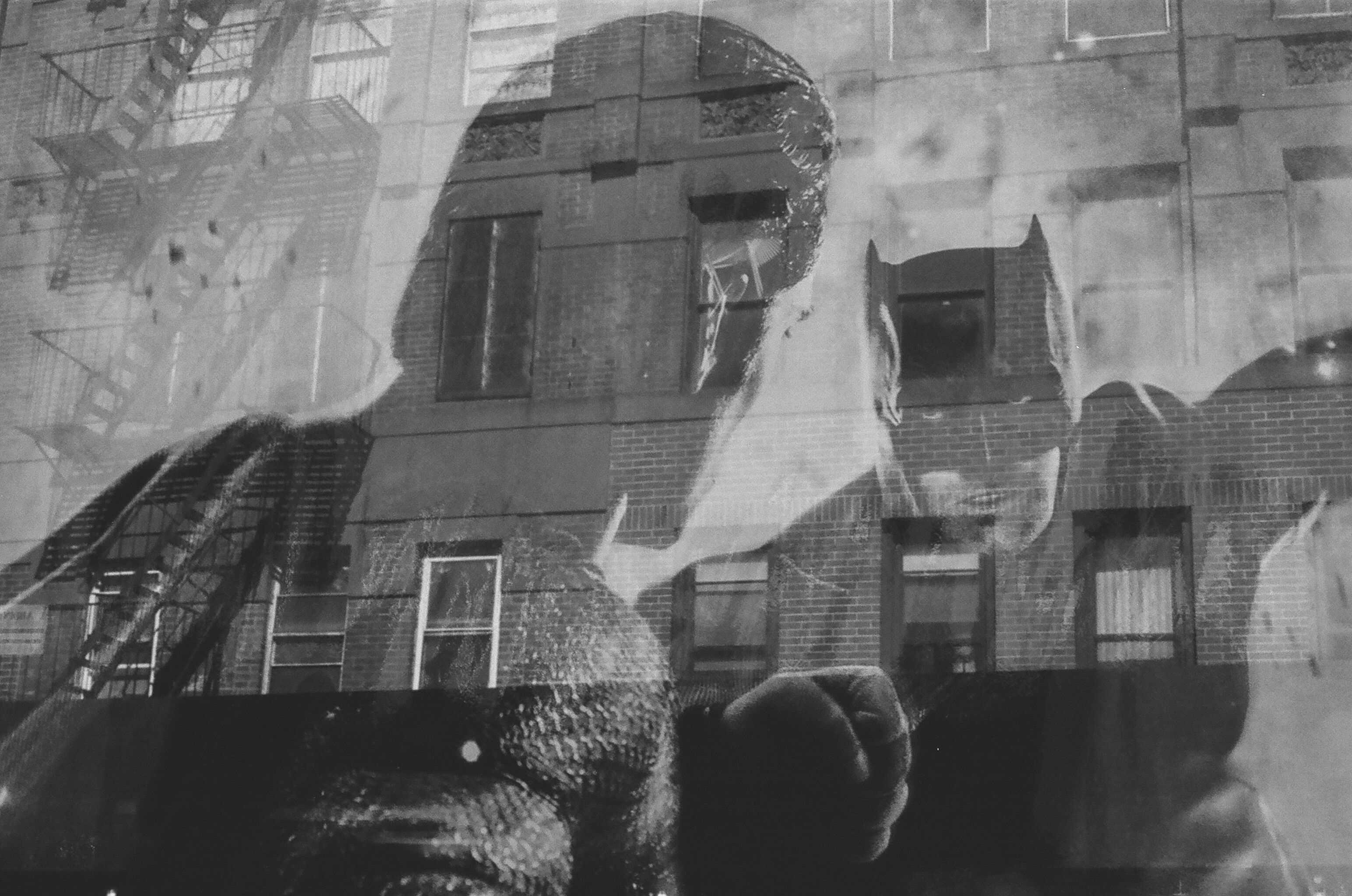 A black-and-white image of Batman fighting another man facing away from the camera overlaid with a translucent façade of walkup apartment buildings in Manhattan