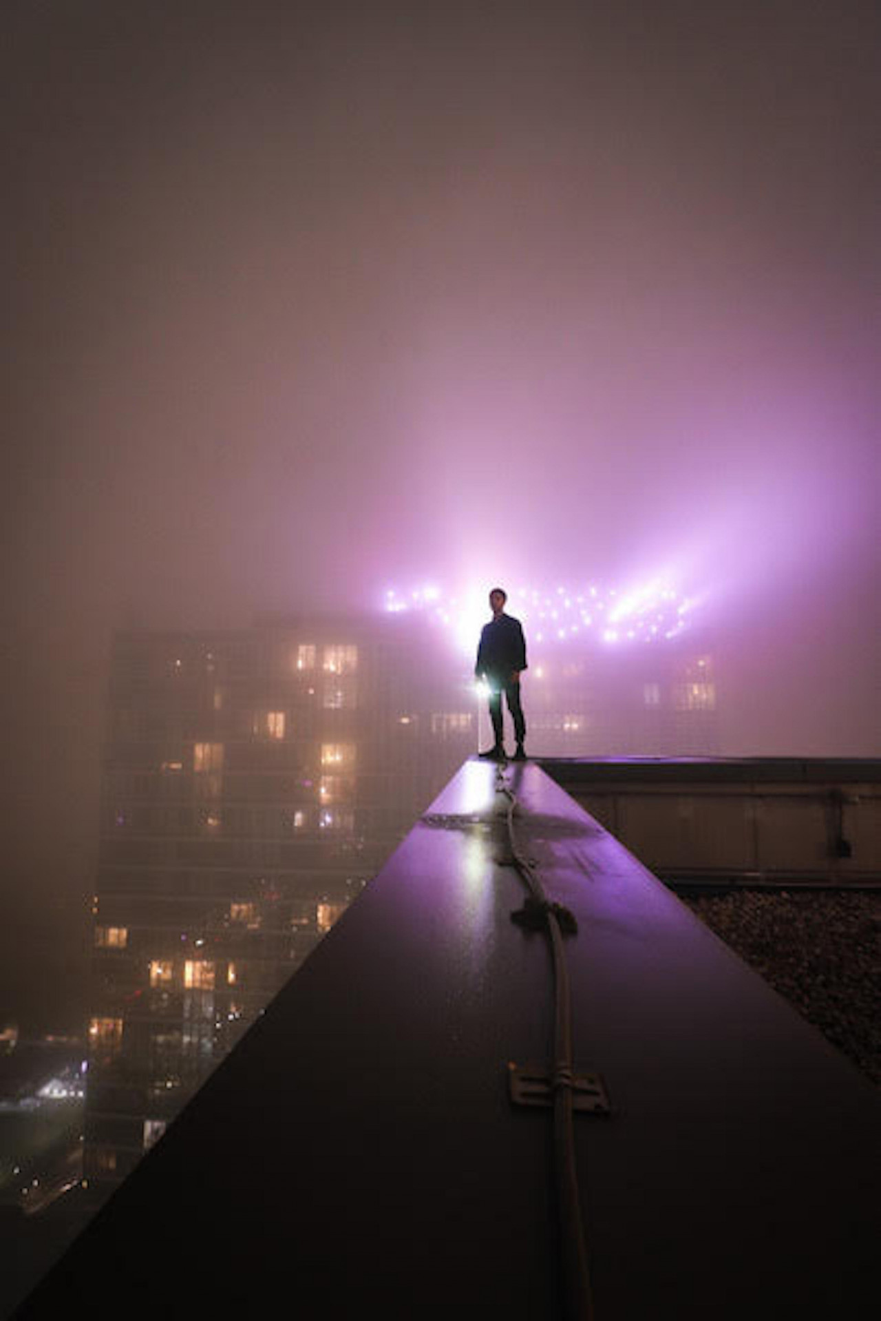 A photograph of a person standing at the corner of a rooftop looking towards the camera. They are surrounded by a light fog, which partially obscures the glow of purple lights and yellow windows of a building behind the figure.