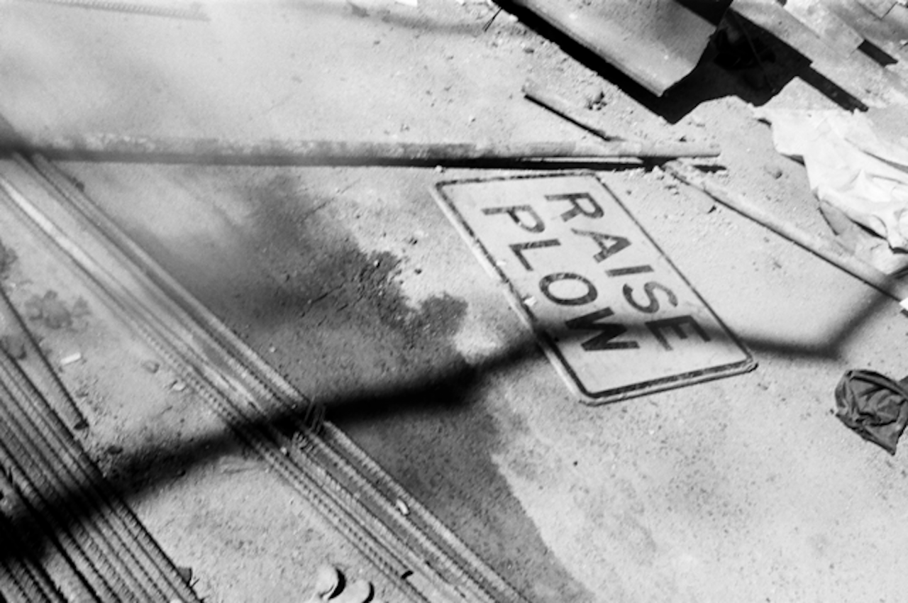 A photograph of a construction site, with several steel beams and a sign titled "RAISE PLOW" lying on the ground