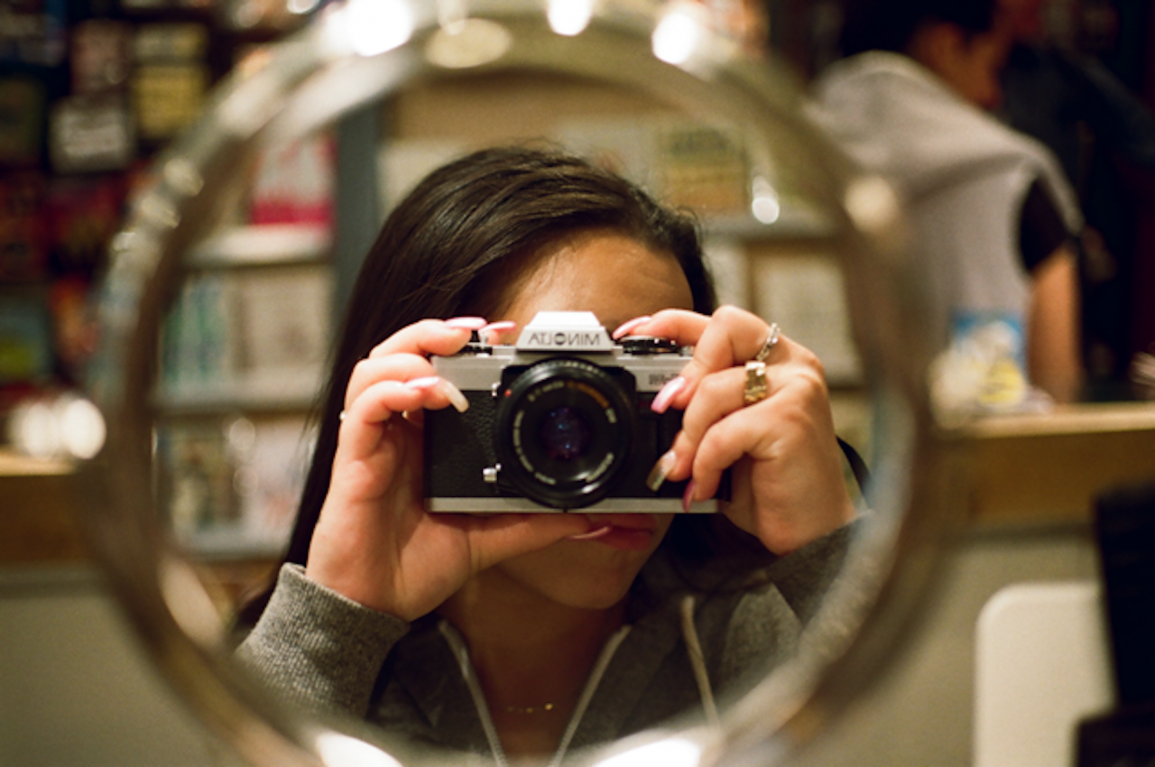 A teenage girl holds a film camera obscuring her face, and takes a photo of her reflection in a small, round mirror. The background is blurred but suggests a retail environment.