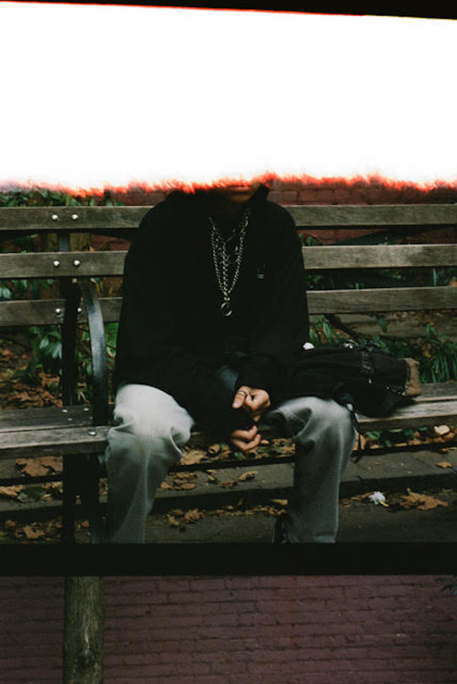 A photograph of  a person with a black hoodie and silver chains sitting on a bench. Their face is hidden by a a swath of white. A band of black borders an image of brick which covers their feet.