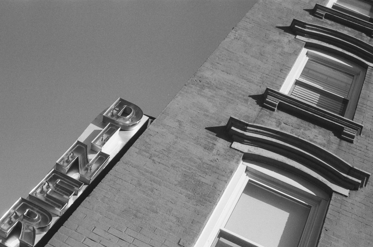 A skewed black-and-white image looking up at the façade of a building and the sky. An LED sign spelling "DINER" hangs from the left side of the building