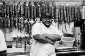 A black-and-white photo of a man smiling with his arms crossed, standing in front of a number of hanging salamis, and wearing a hat with a logo stating 