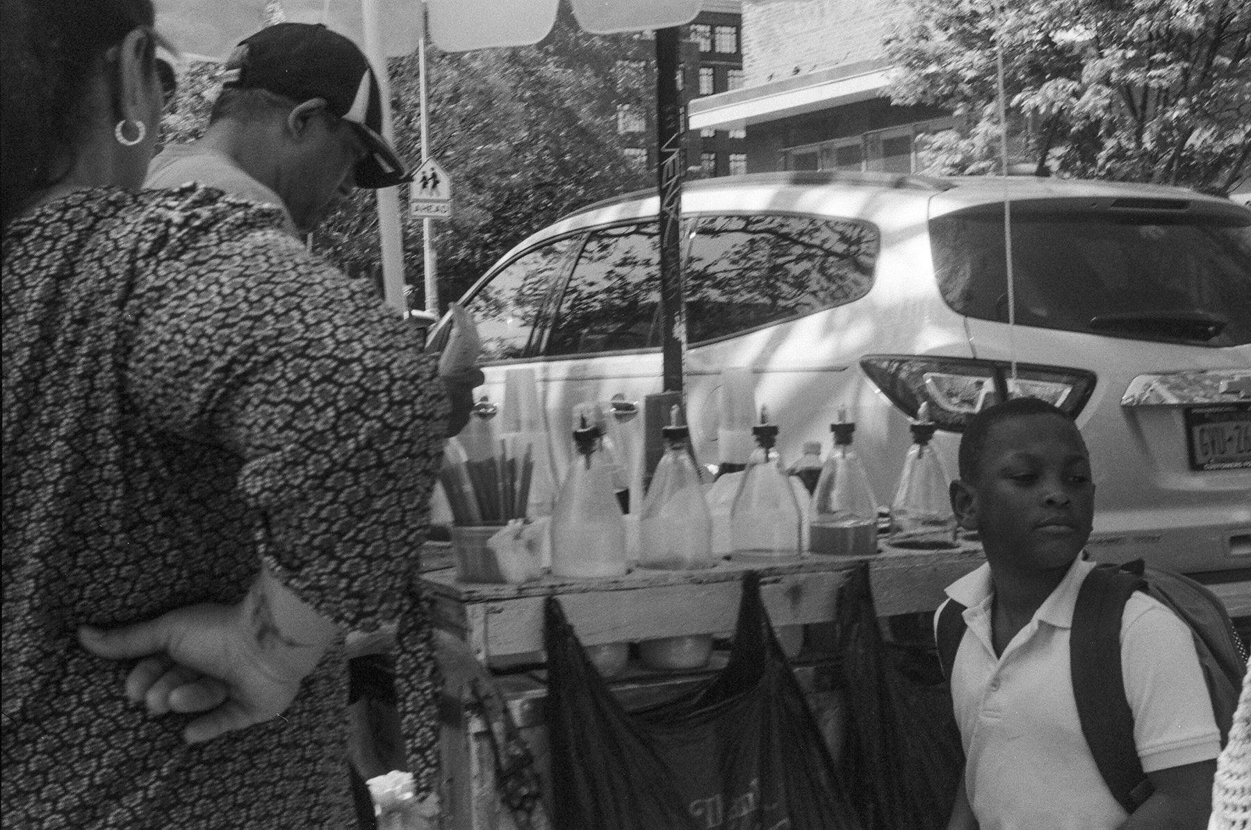 A boy and two adults standing around an italian ice street cart in front of an SUV.