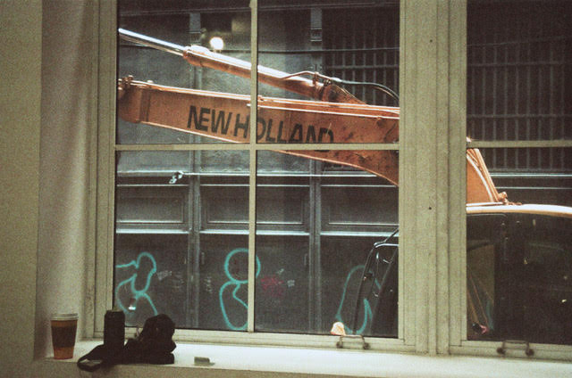 A window sill with cups, cans and cloth is visible and a crane can be seen outside of the window with the text: "New Holland" written on it. Circular green graffitti is visible on the facade of the building behind the crane.