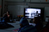 A photo of students sitting around a table, watching a YouTube video and speaking to one another.