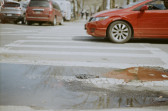 A photo of a crosswalk with a red car standing halfway in it, and a small puddle of water in the foreground.