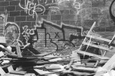 A black-and-white photo of a brick wall covered in graffiti and a pile of broken wood, on top of a crumpled sign stating 