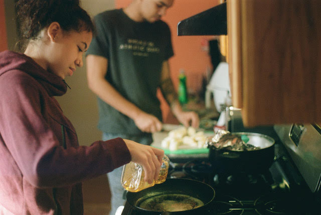 A figure wearing a red hoodie pours oil into a cooking pan on a stove while a figure wearing a T-shirt and jeans in the background chops a produce item. Another pot is visible on the stove along and a cabinet and light appear above the stove.
