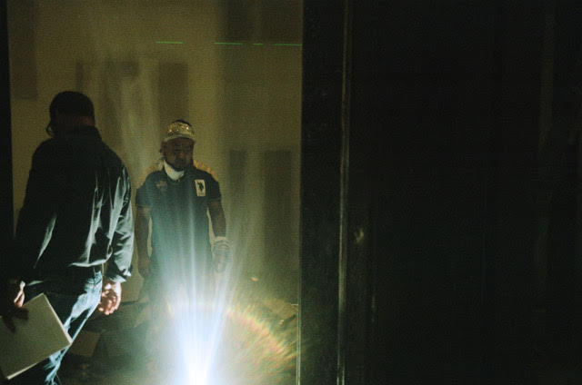 One figure is looking at the ground with their back facing the camera and another painted figure appears frontally looking down. They both are around a bright light emerging from below. A black wall covers the right half of the image.