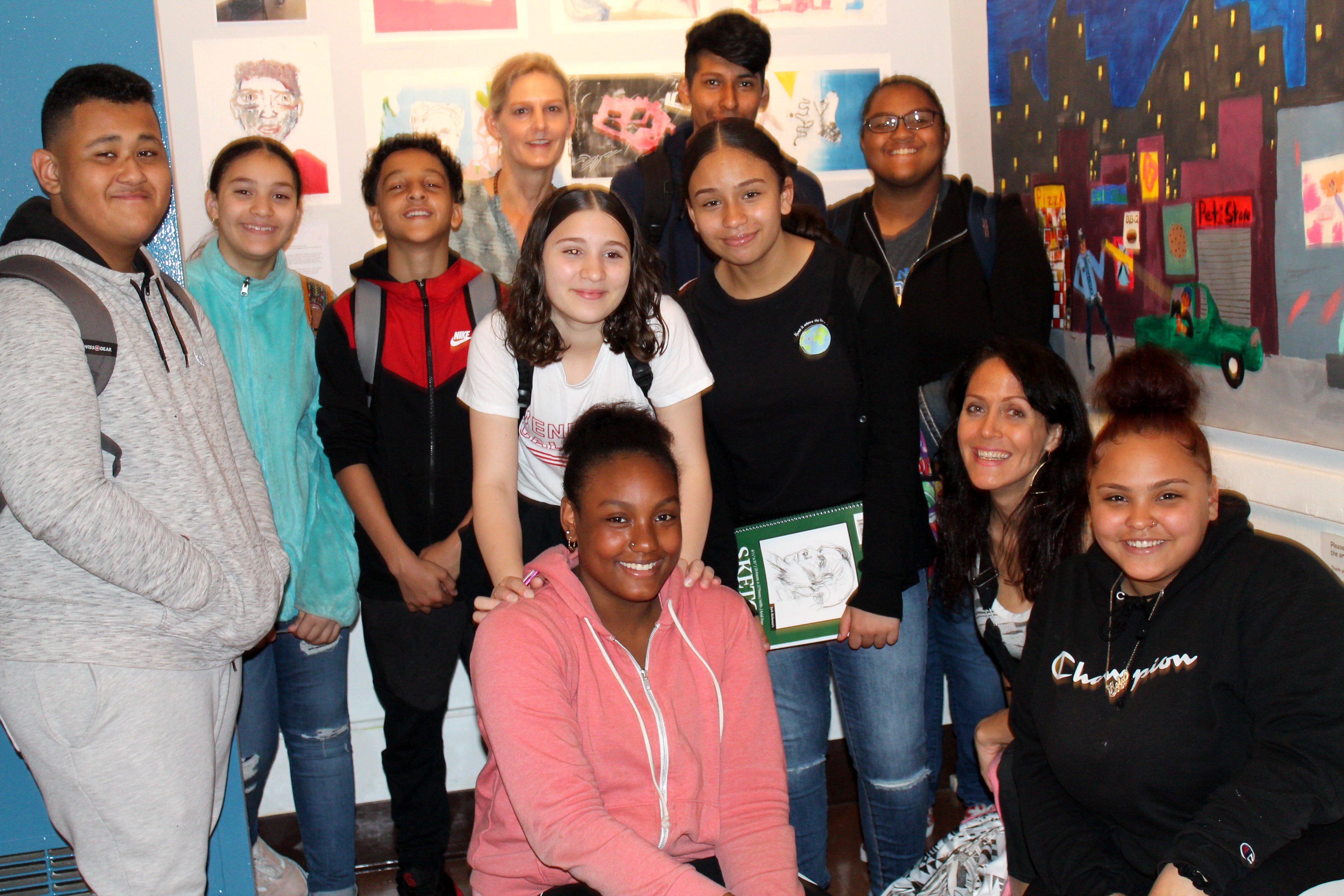 Several students and two teachers smiling for the camera in front of their artwork hanging on the wall
