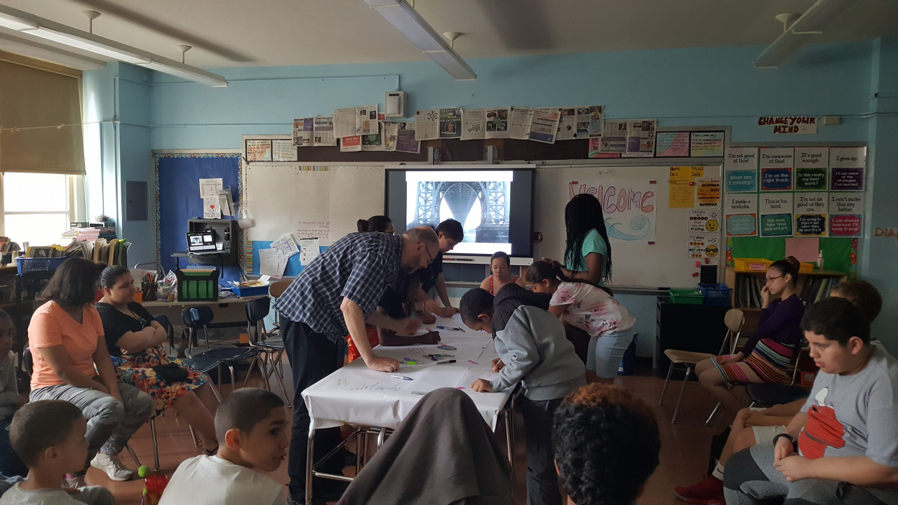 A photograph of a classroom environment with several students sitting around the edges of the room and a few drawing directly onto a white tablecloth in the center of the room. An image of the underside of a large bridge is shown on a monitor at the front of the room