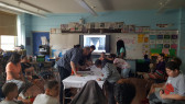 A photograph of a classroom environment with several students sitting around the edges of the room and a few drawing directly onto a white tablecloth in the center of the room. An image of the underside of a large bridge is shown on a monitor at the front of the room