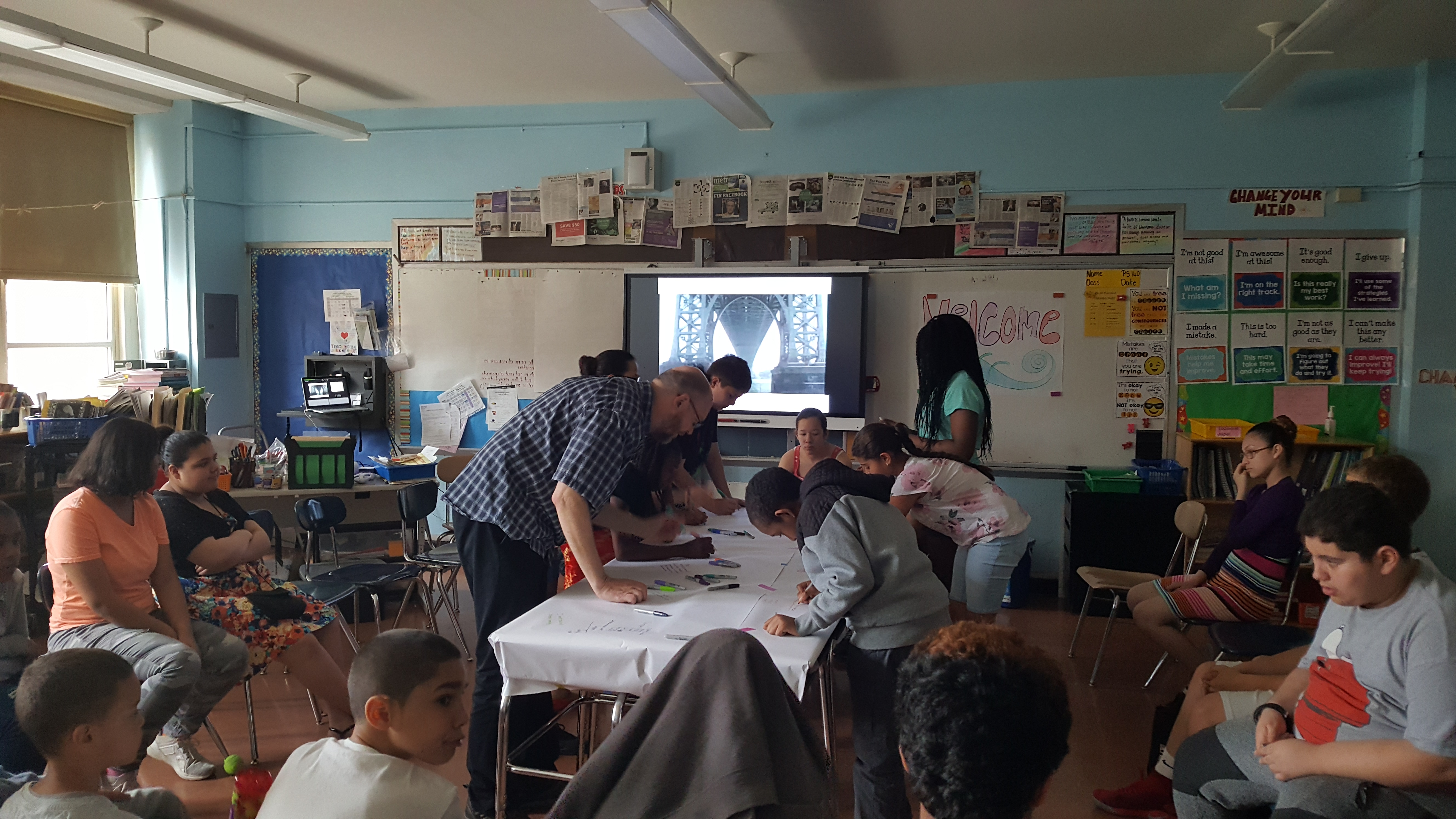 A photograph of a classroom environment with several students sitting around the edges of the room and a few drawing directly onto a white tablecloth in the center of the room. An image of the underside of a large bridge is shown on a monitor at the front of the room