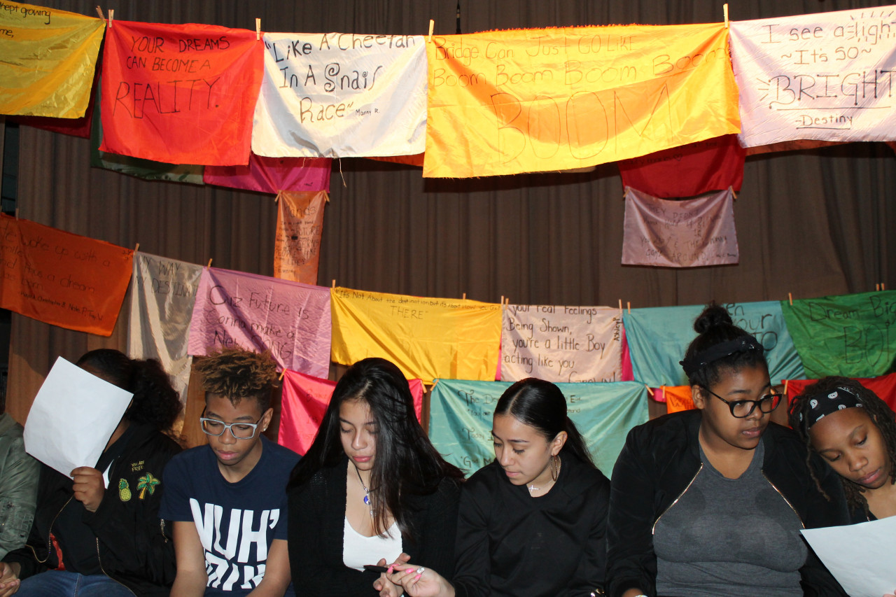 Several students sit, looking down towards printed sheets of paper, in front of colorful silk banners with writing stating "Your dreams can become a reality," "like a cheetah in a snail
