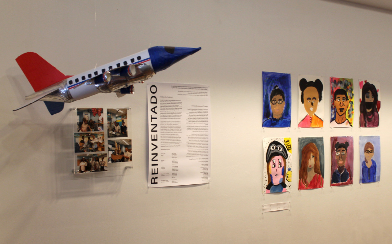 An exhibition view of various artworks. Several stylized portraits hang on the right; in the middle, a large didactic titled "REINVENTADO." On the left, several small photos of students in a classroom hang underneath a red-white-and blue airplane suspended from the ceiling