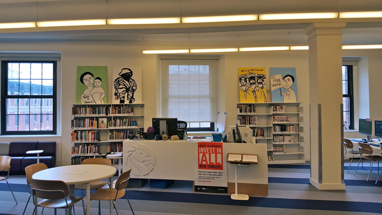 A photo of a front desk in a library - several desks with computers, bookshelves, and a couch are also visible. Four paintings lie on top of the bookshelves, depicting stylized figures against block color backgrounds