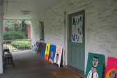 A photo of the exterior of a building, with several colorful portrait paintings lying on the wooden porch. One has text stating 