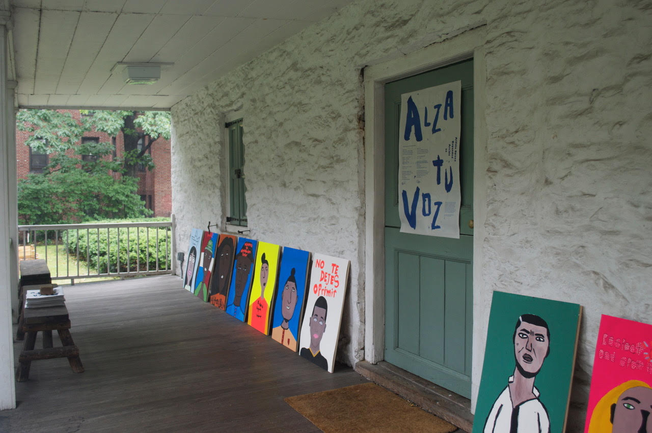 A photo of the exterior of a building, with several colorful portrait paintings lying on the wooden porch. One has text stating "no te dejes oprimir." A large poster hangs on the door to the building, with the title "Alza tu Voz"