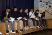 Several students sit on the edge of a wooden stage holding printed sheets of paper. One girl reads from her paper while holding a microphone