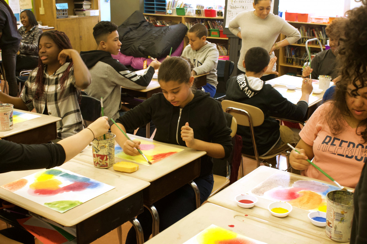 A classroom with several students, each at their own desk, painting with primary-color watercolors
