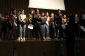 A photograph of a group of students standing onstage, holding printed paper, and many smiling